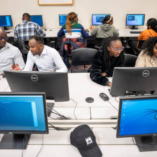 A classroom setting shows students working at desktop computers featuring the Kali Linux operating system. The students are arranged in pairs or groups, engaged in collaborative work, with a black baseball cap visible on one of the front tables.